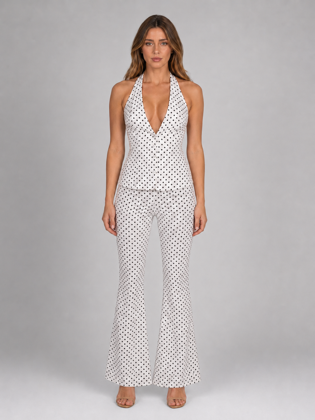 Woman wearing white halter polka dot jumpsuit with wide leg trousers and tan sandals against gray background