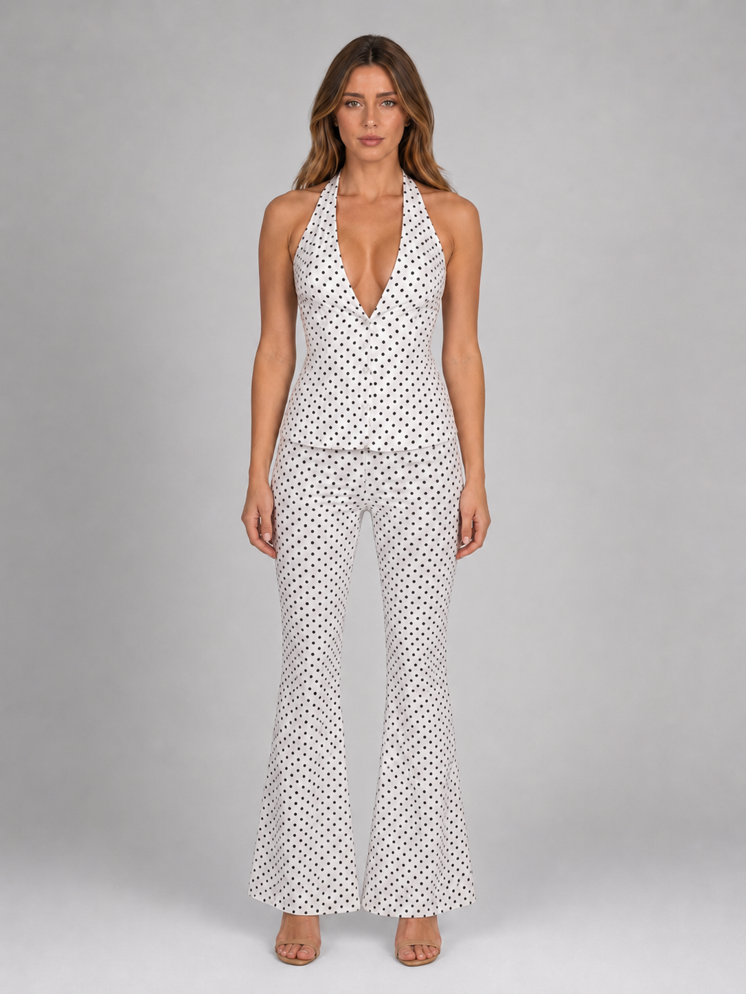 Woman wearing white halter polka dot jumpsuit with wide leg trousers and tan sandals against gray background