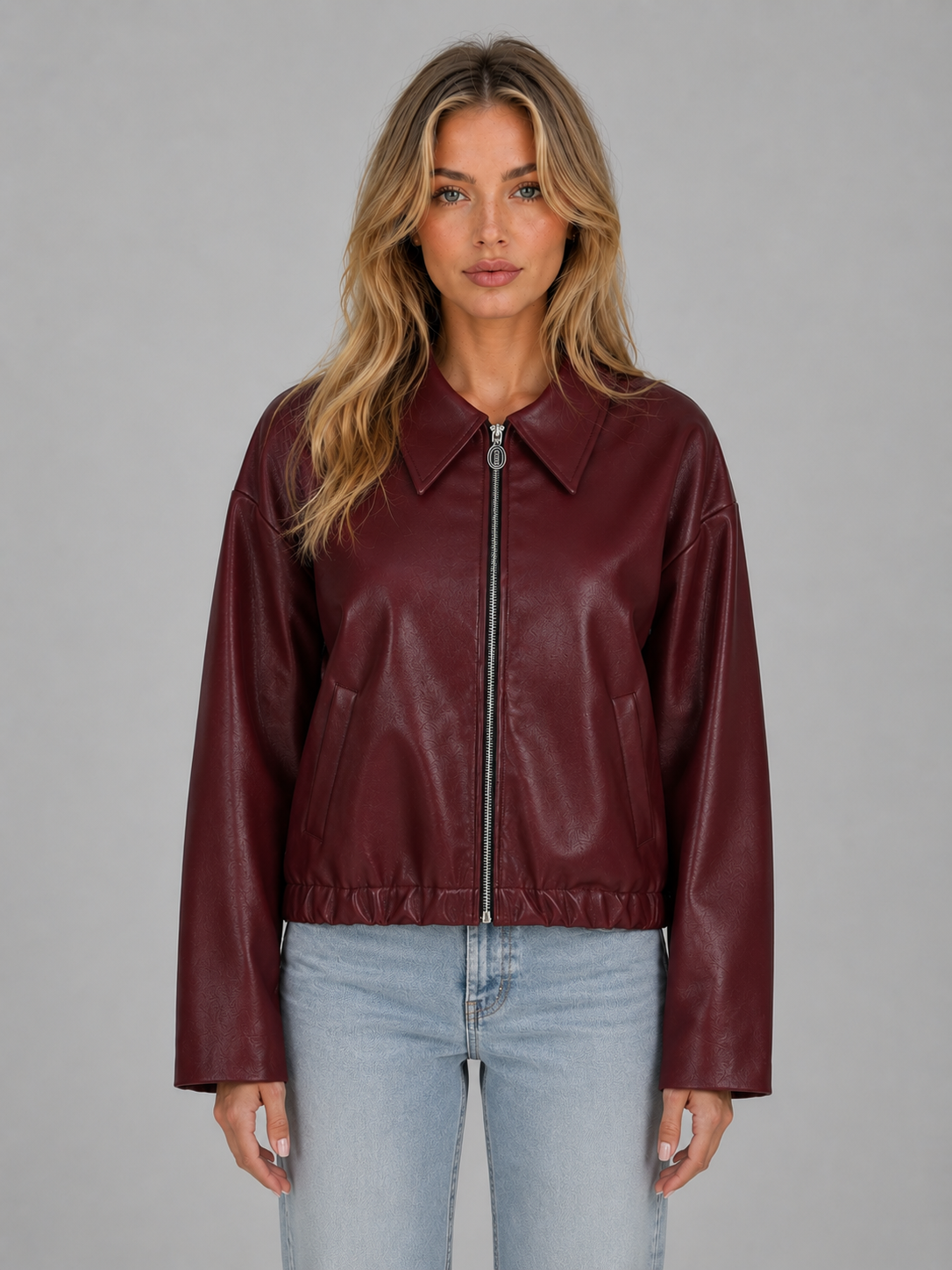 Woman wearing burgundy cropped leather zip jacket with collar and pockets over light blue jeans against gray background
