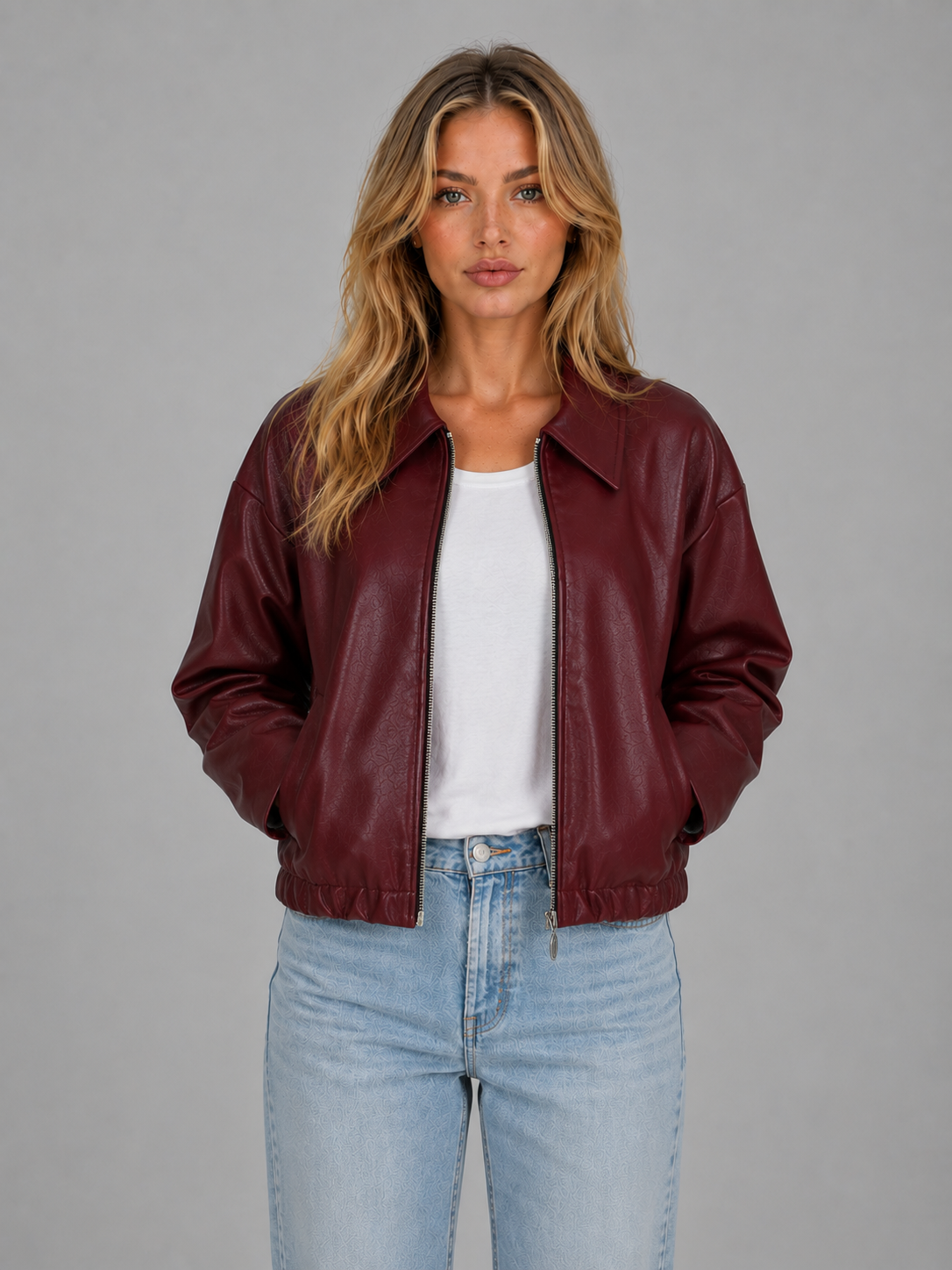 Woman wearing burgundy cropped leather zip jacket with white shirt and light blue jeans against gray background