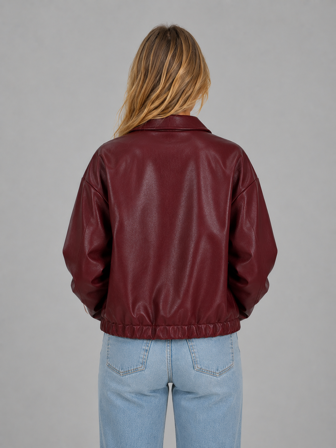 Woman wearing burgundy cropped leather jacket with collar and pockets over light blue jeans, back view
