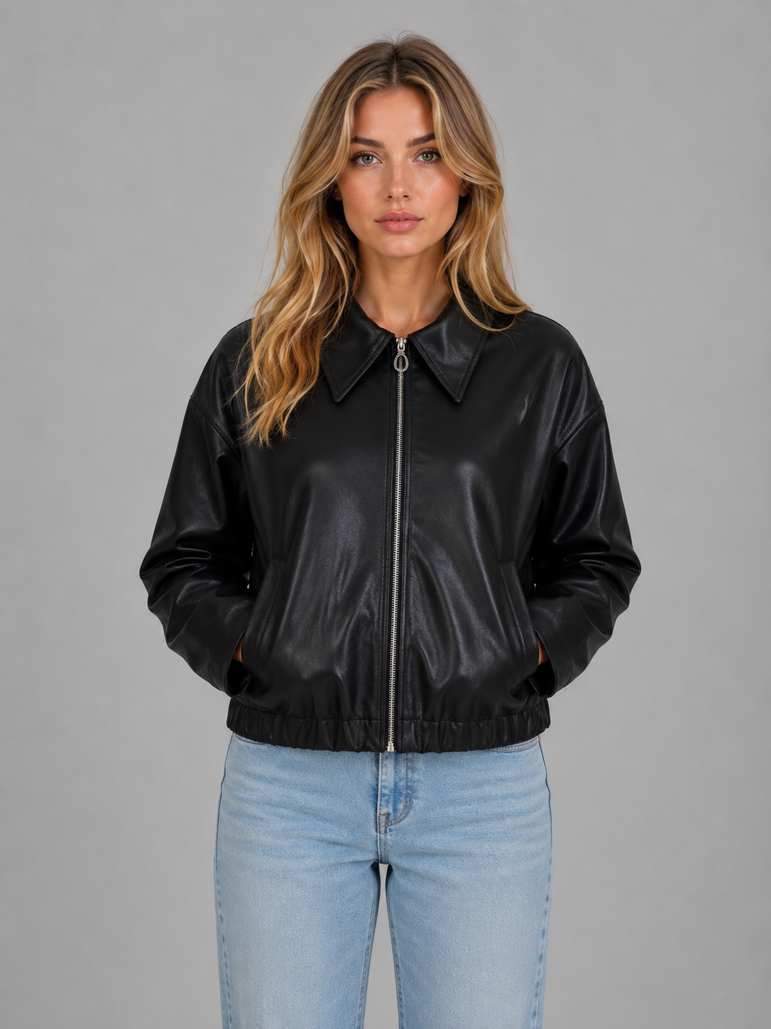 Woman wearing black leather cropped zip jacket with collar and pockets, styled with light blue jeans against gray background