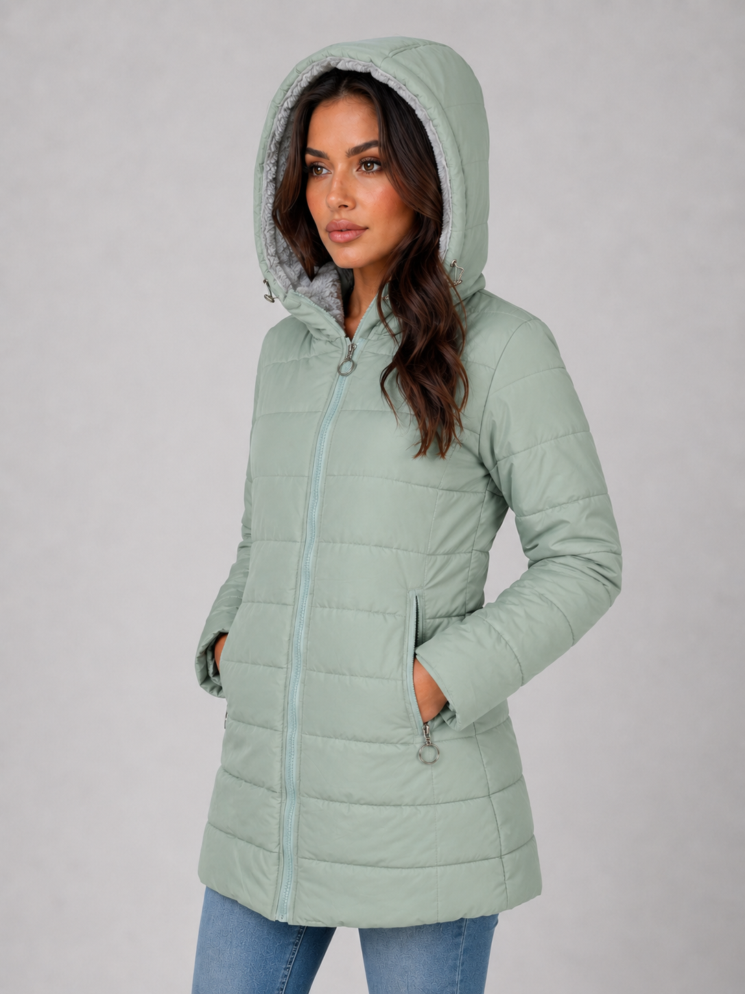 Woman wearing sage green hooded quilted puffer jacket with fleece lining and front zip closure, styled with blue jeans