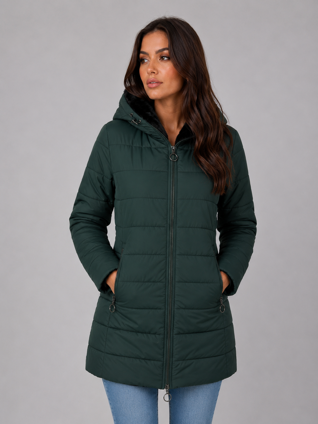 Woman wearing dark green hooded quilted puffer jacket with fleece lining over light blue jeans, front view against gray background