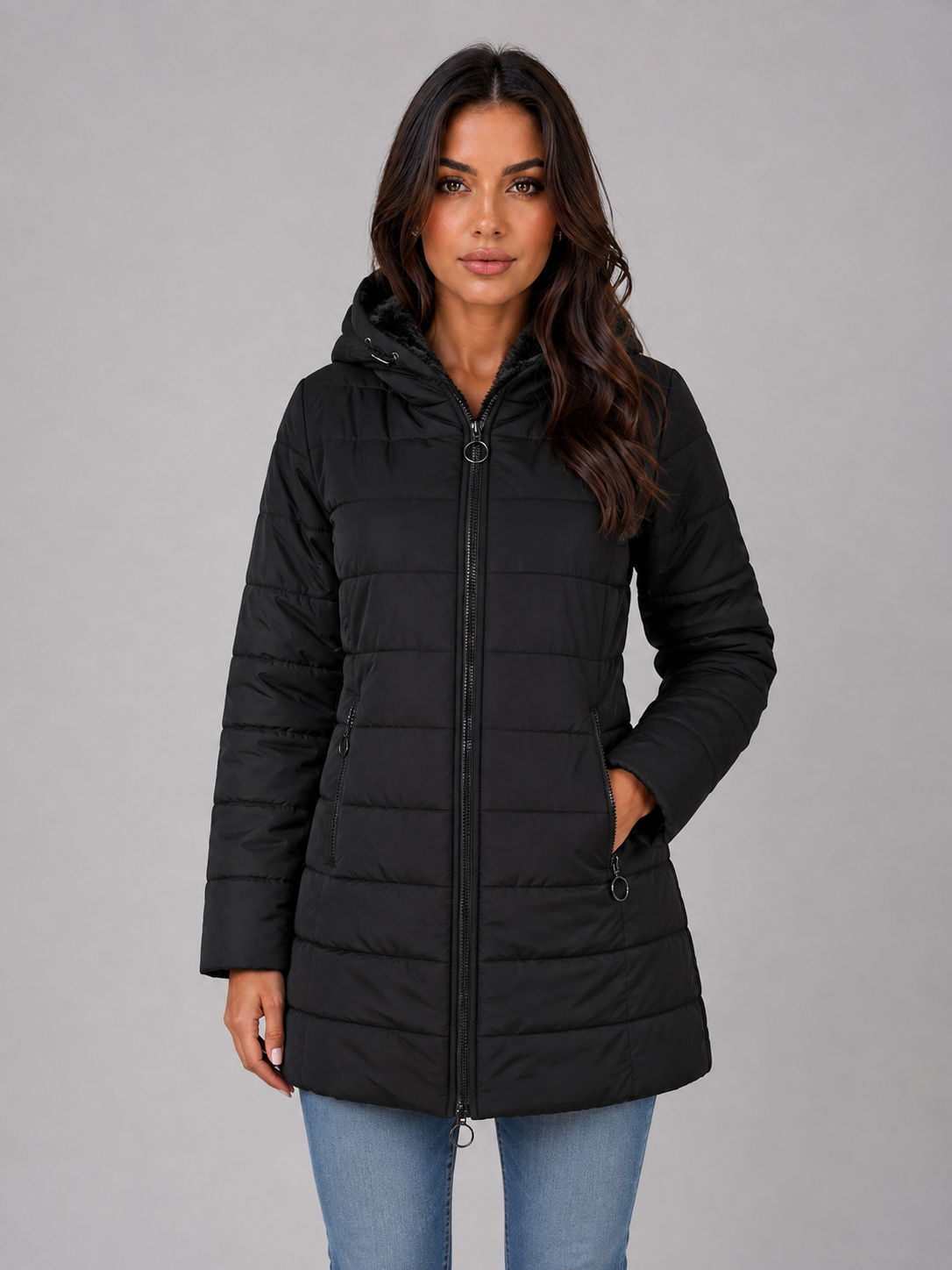 Woman wearing black hooded quilted puffer winter jacket with fleece lining, paired with light blue jeans against gray background