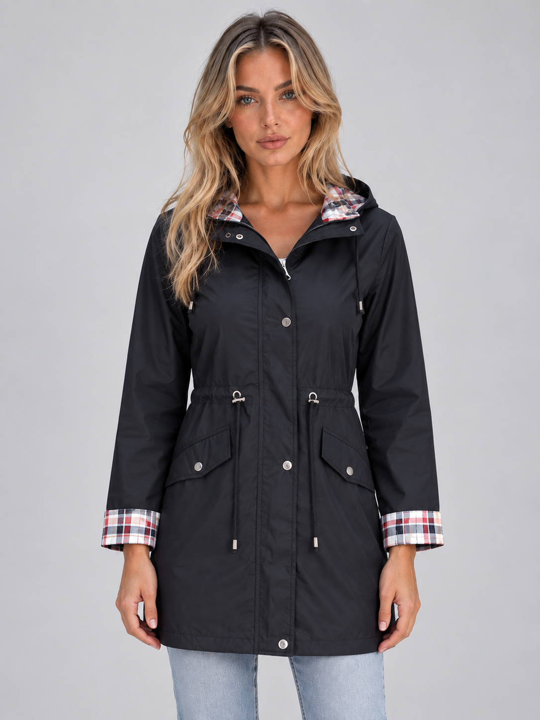 Women's black zip-up hooded raincoat parka with plaid lining, drawstring waist, and chest pockets worn over light blue jeans
