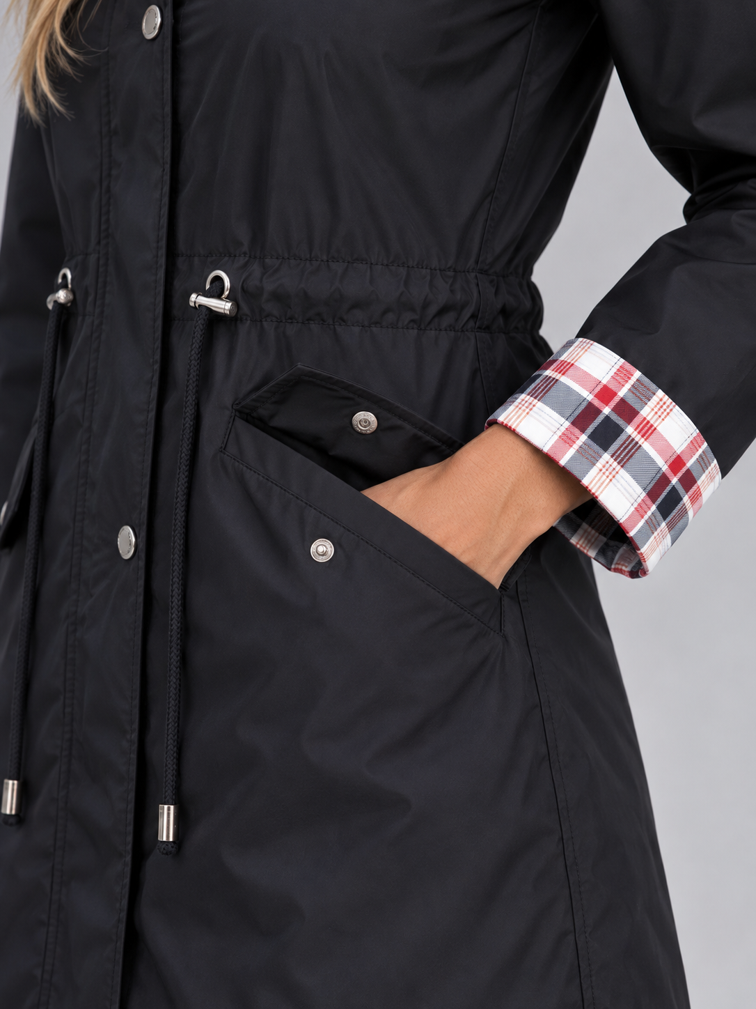 Black raincoat parka with plaid shirt cuff visible, showing drawstring waist, front pockets, and silver toggle details
