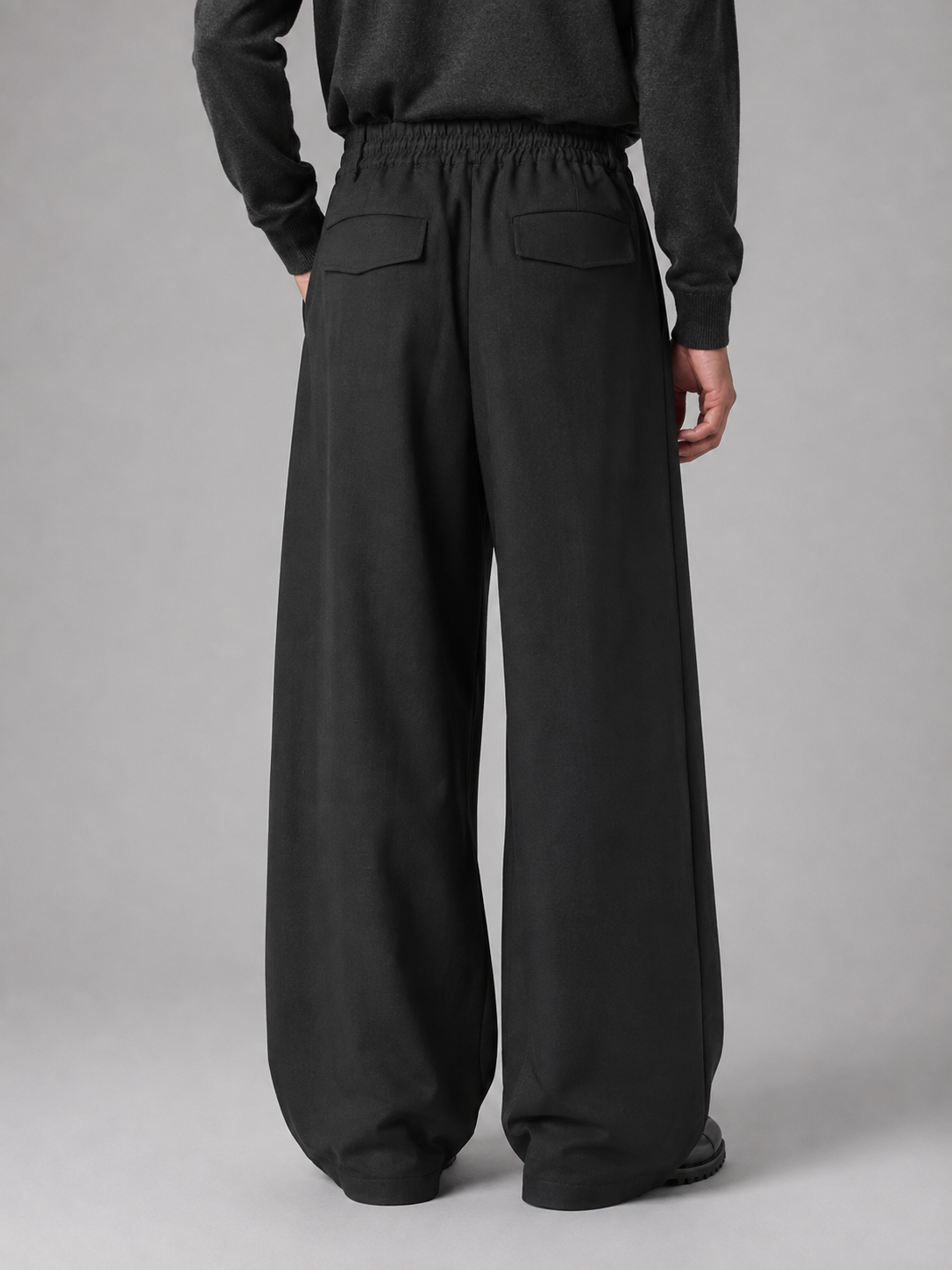 Men's black pleated baggy wide leg trousers with elastic waist and rear patch pockets, back view