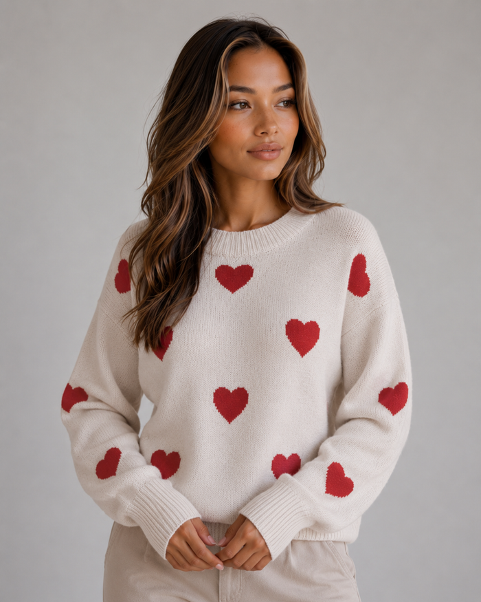 Woman wearing cream crew neck knitted sweater with red heart pattern scattered across front and sleeves