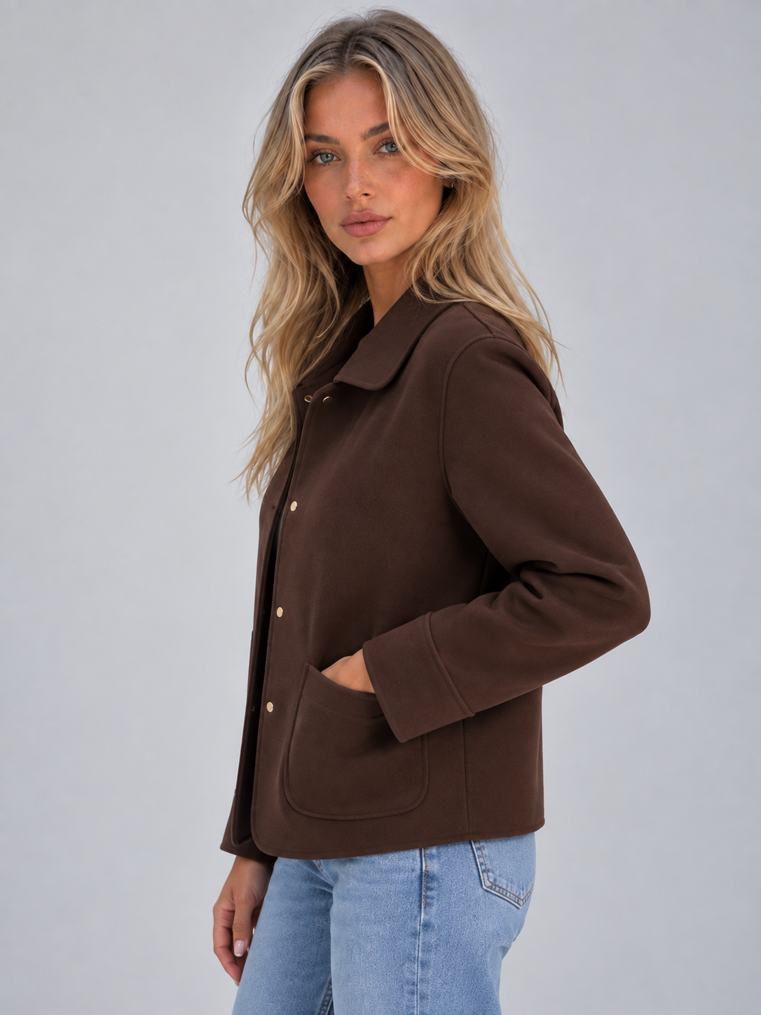 Woman wearing brown collared button-up overshirt jacket with patch pockets and gold buttons, styled with light blue jeans