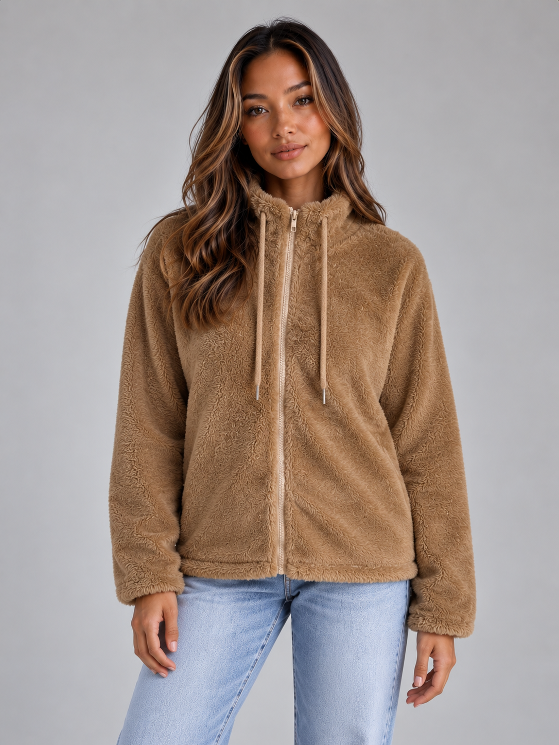 Woman wearing tan teddy fleece zip-up jacket with hoodie over light blue jeans against gray background