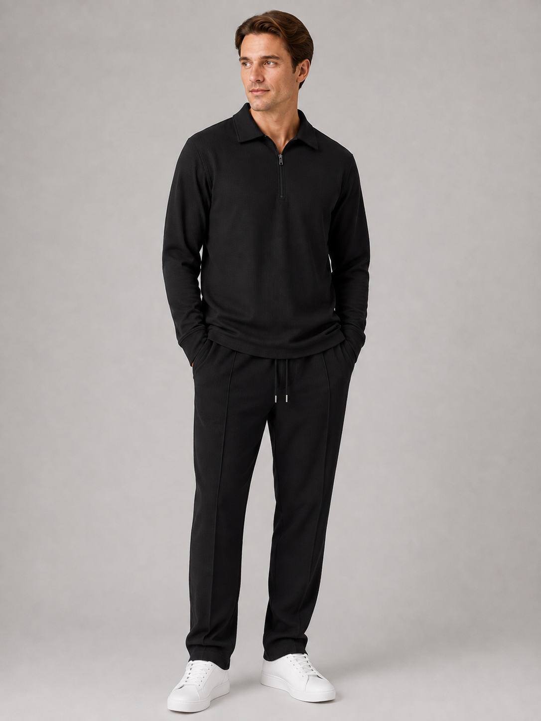 Man wearing black quarter-zip polo shirt and black textured trousers with white sneakers, standing against gray background