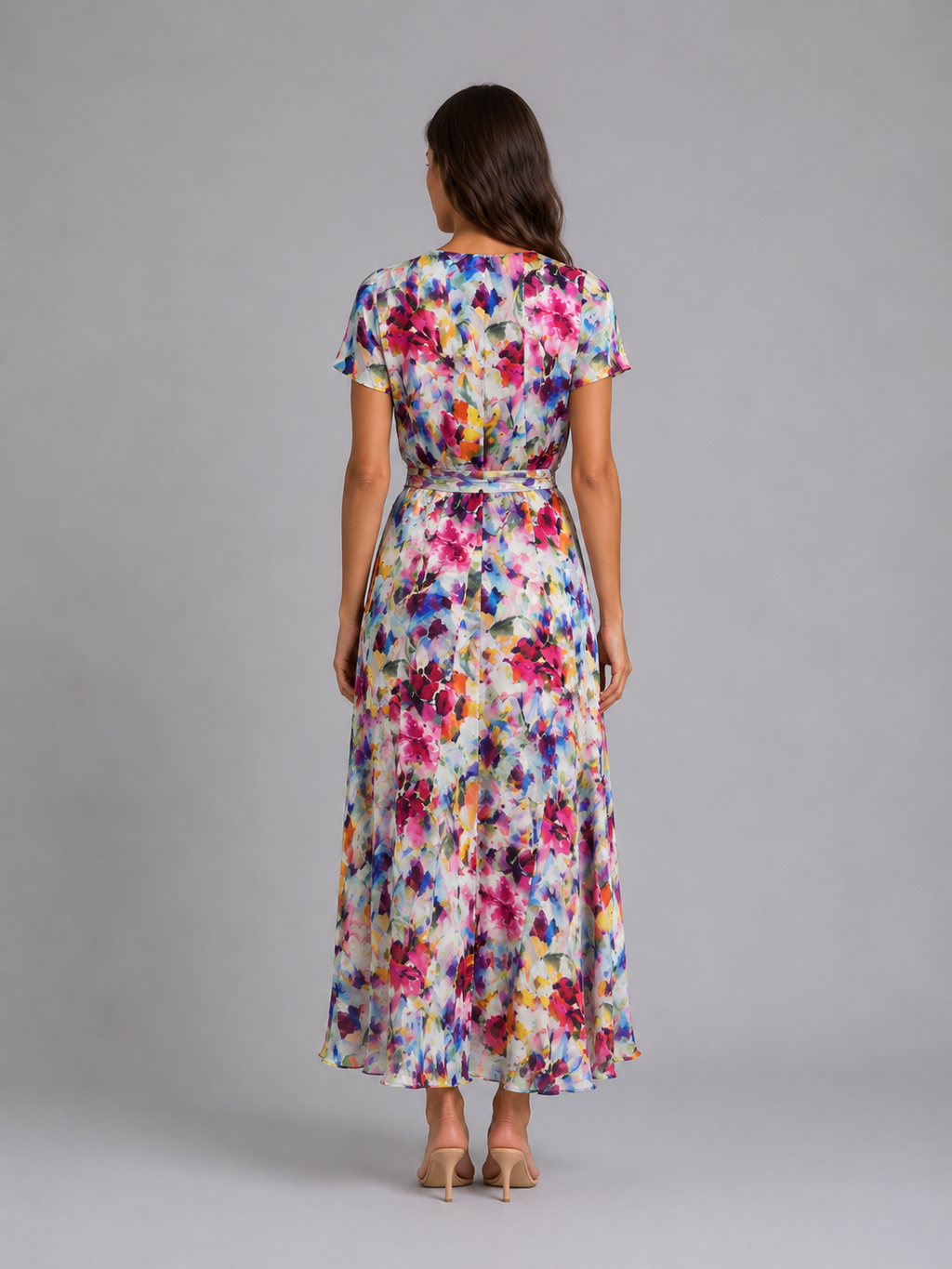 Woman wearing a colorful floral midi dress with short sleeves and tie waist, viewed from behind, standing against a gray background