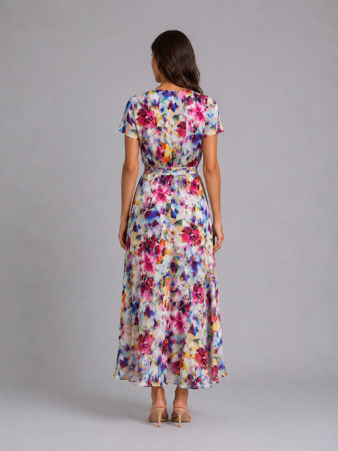 Woman wearing a colorful floral midi dress with short sleeves and tie waist, viewed from behind, standing against a gray background