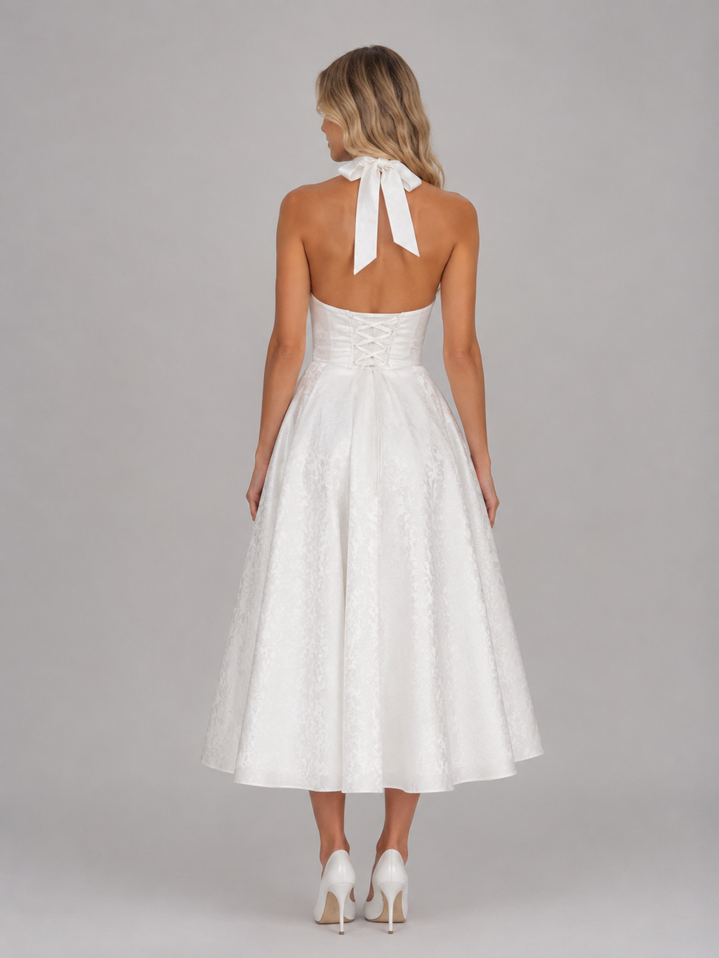 Woman wearing white jacquard halter midi dress with tied neckline, corset back, and full skirt, styled with white heels