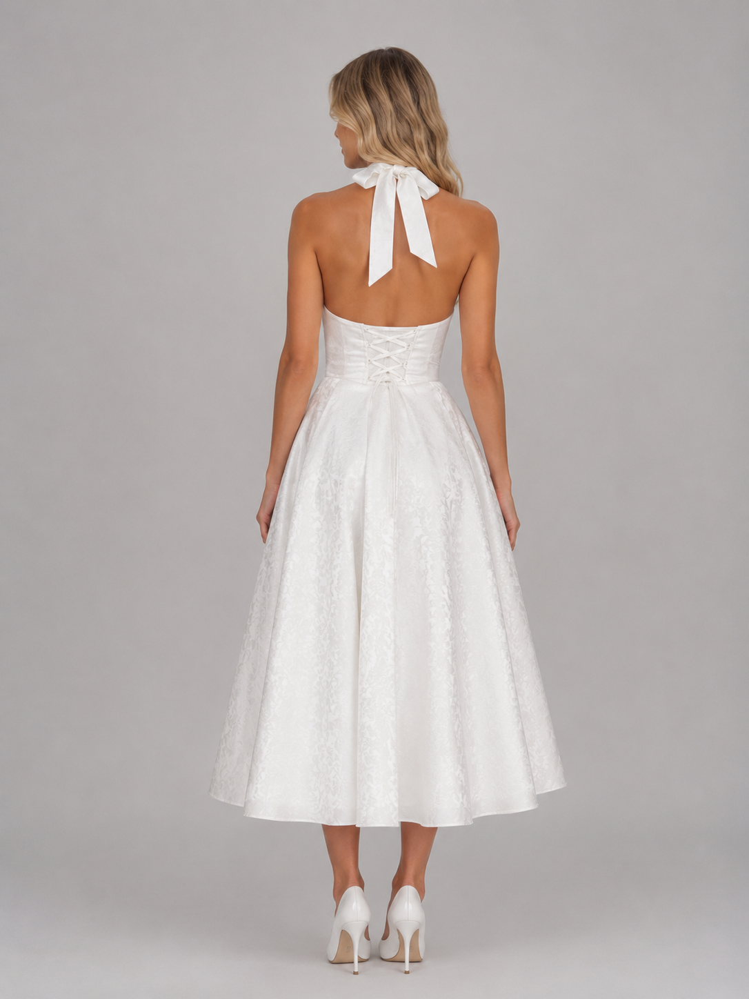 Woman wearing white jacquard halter midi dress with tied neckline, corset back, and full skirt, styled with white heels