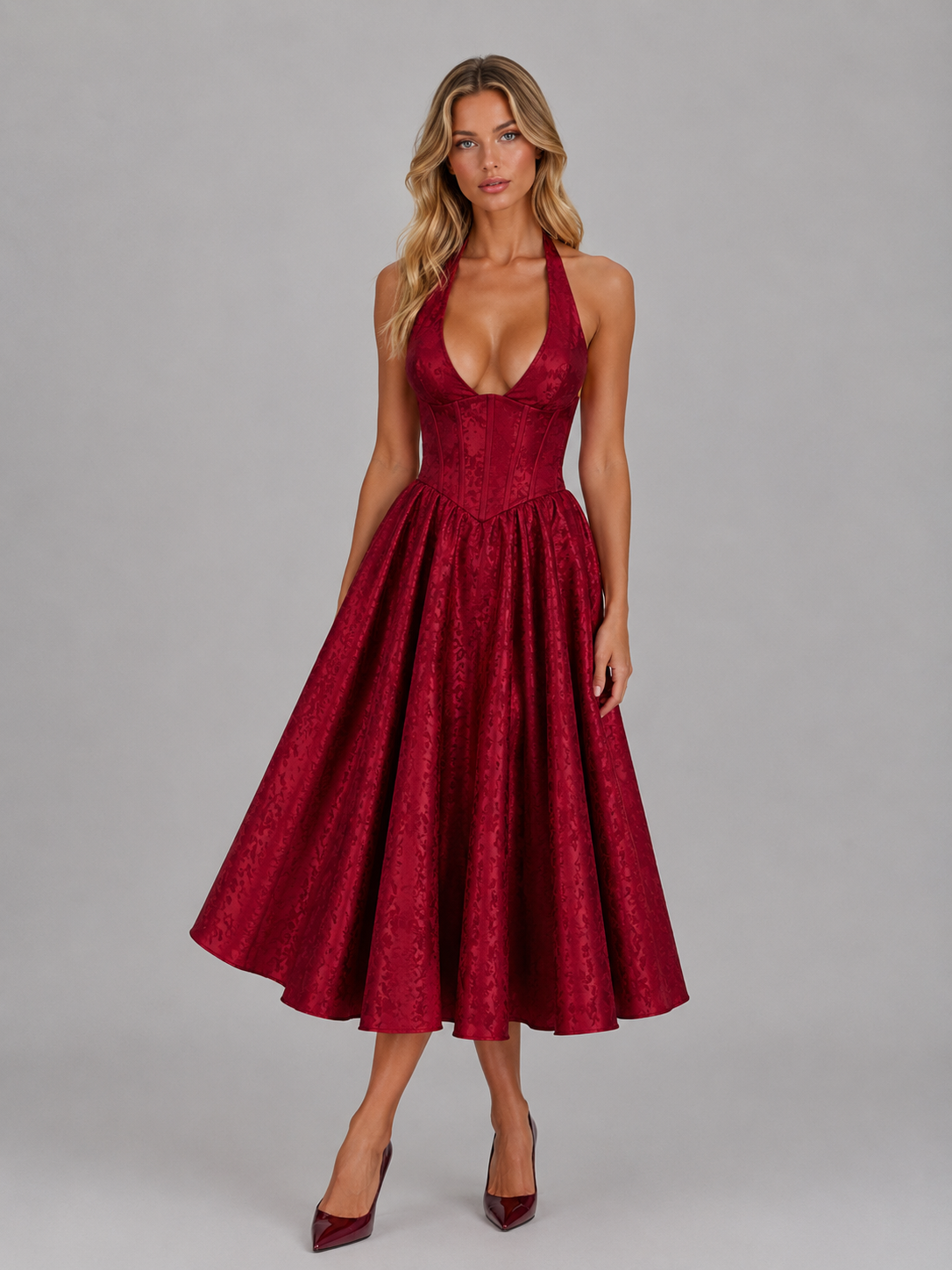 Woman wearing burgundy jacquard halter midi dress with full skirt and matching velvet heels against gray background
