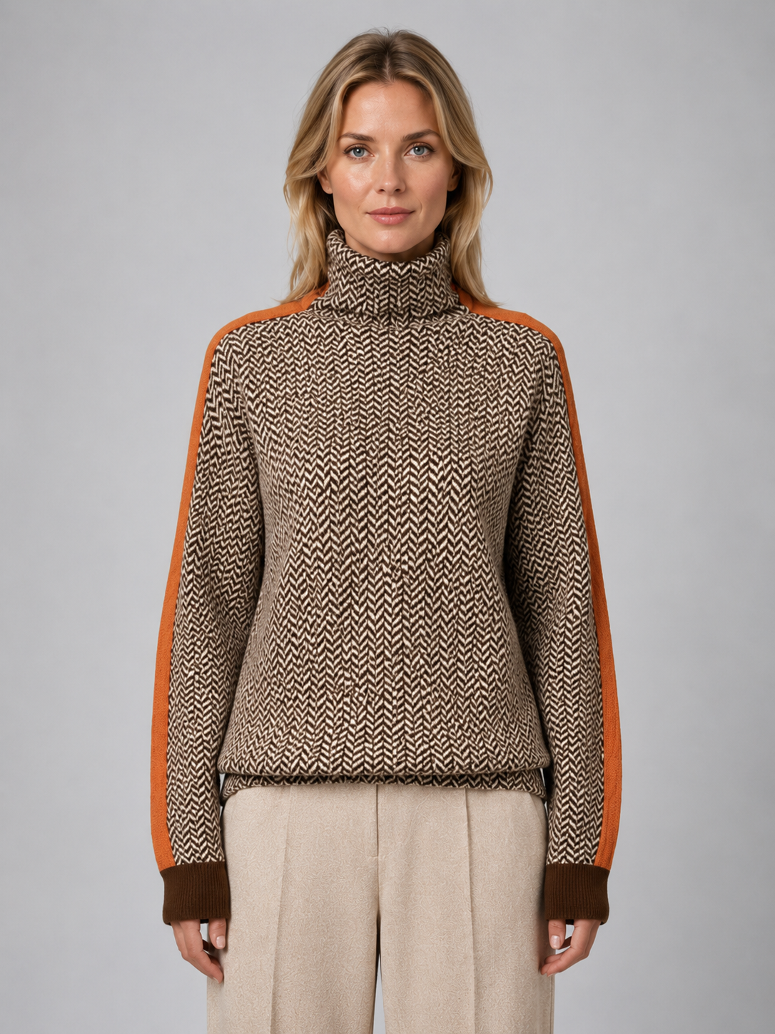 Woman wearing brown herringbone turtleneck jumper with orange sleeves and cream pants against gray background