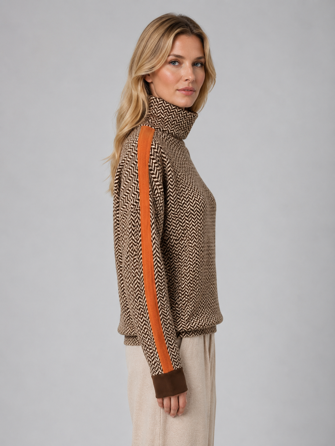 Woman wearing beige herringbone knit turtleneck jumper with orange trim and cream trousers, shown from side profile