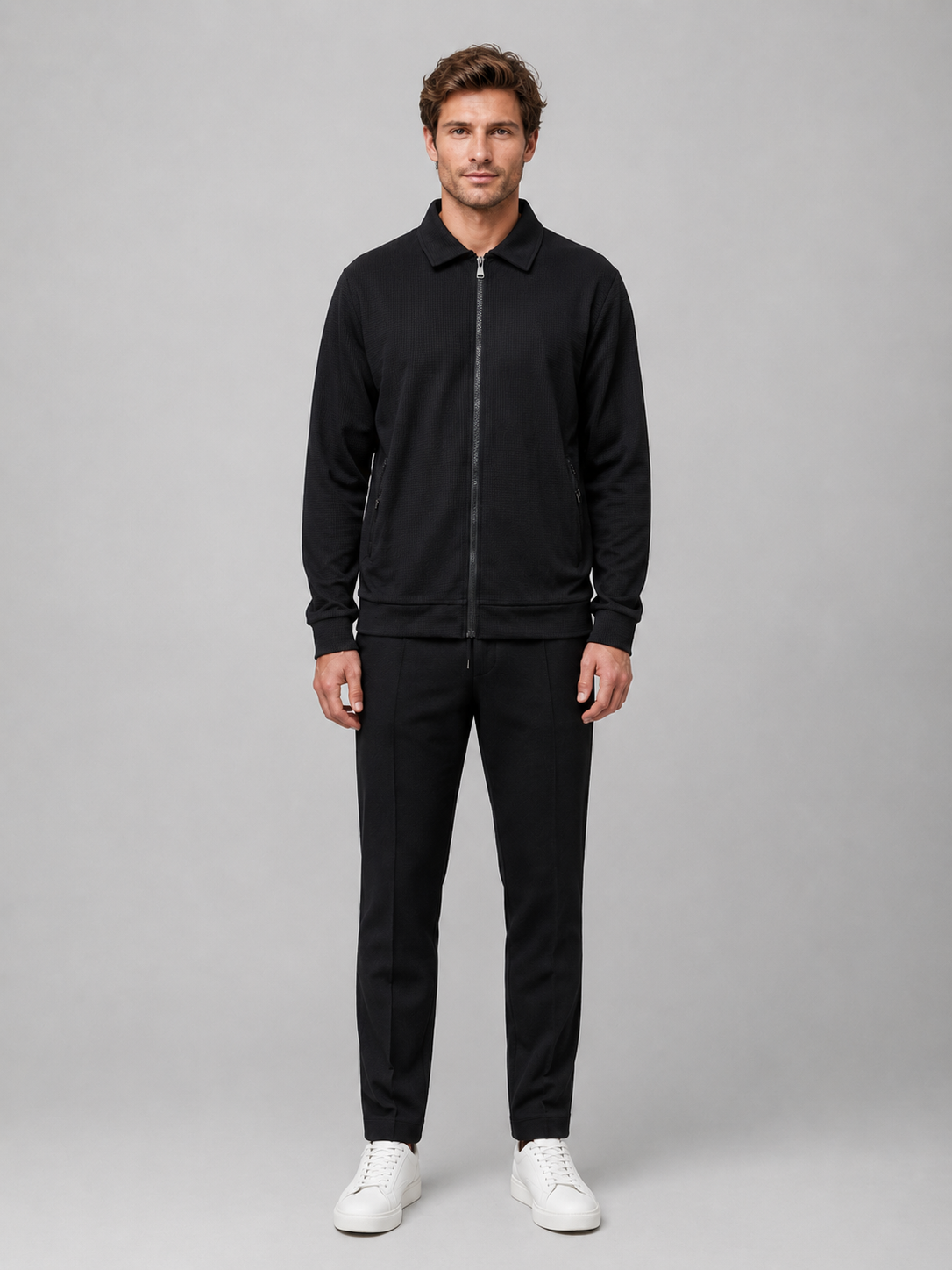 Man wearing black textured zip jacket and black trousers with white sneakers, standing against neutral background