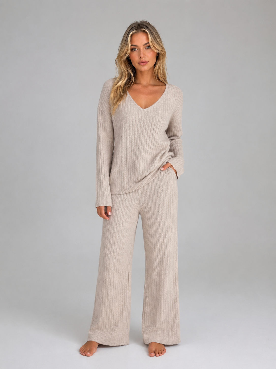 Woman wearing beige ribbed knit V-neck jumper and matching wide leg trousers lounge set, standing against grey background