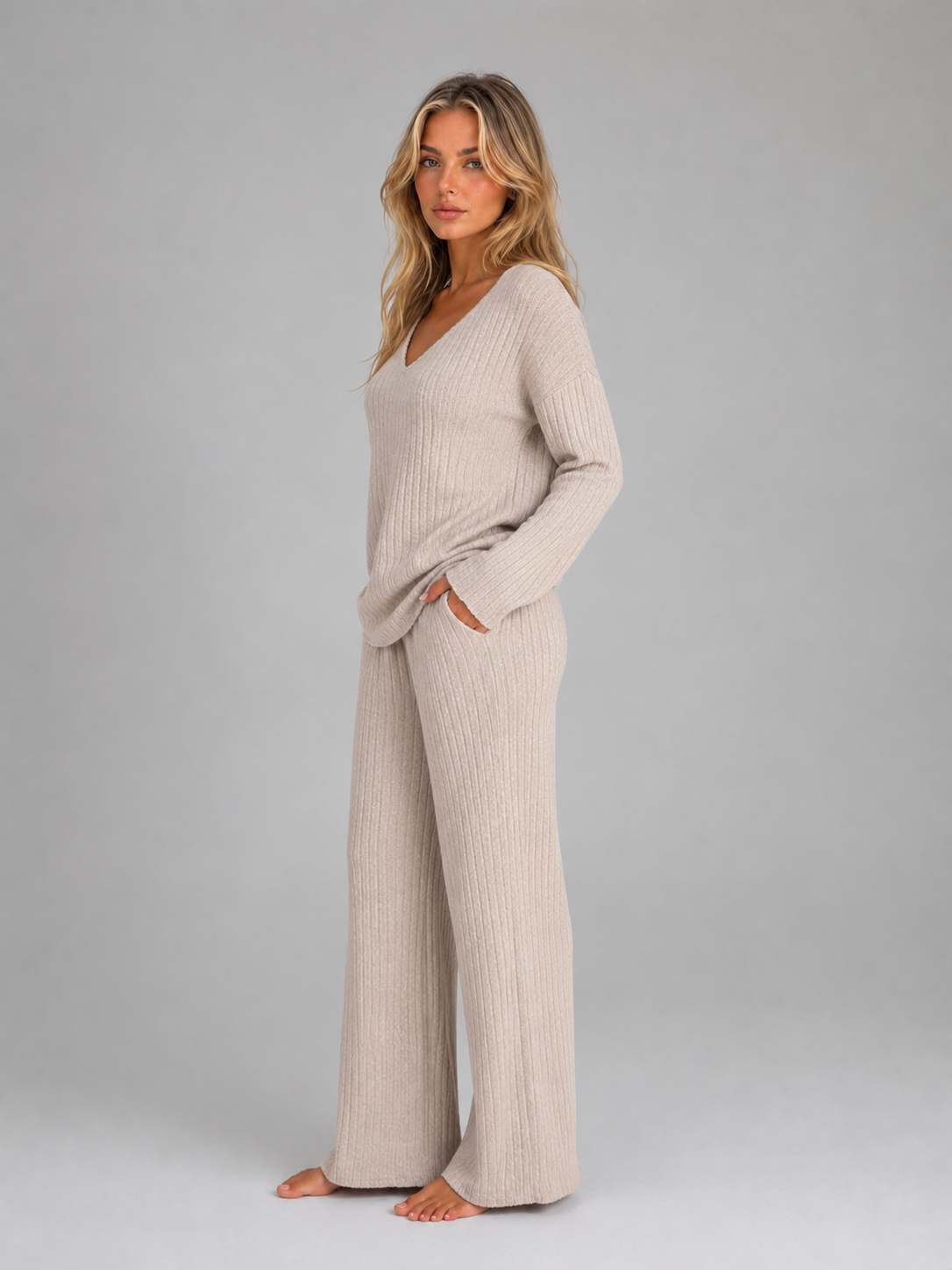 Woman wearing cream ribbed knit v-neck jumper and matching wide leg lounge trousers, barefoot against gray background