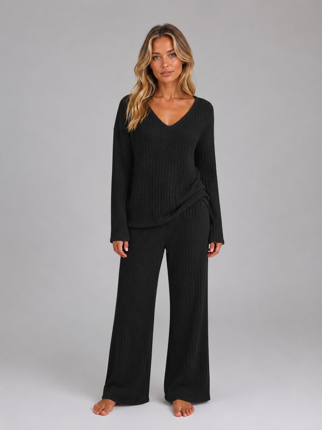 Woman wearing black ribbed knit v-neck jumper and matching wide leg trousers lounge set, barefoot against gray background