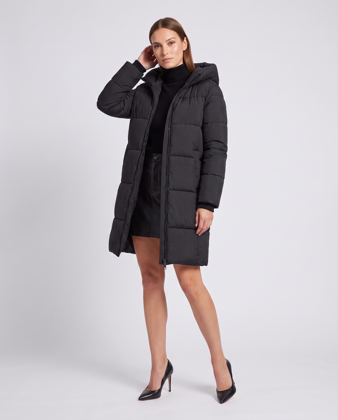 Woman wearing black quilted puffer coat with hood over black turtleneck, paired with black heels