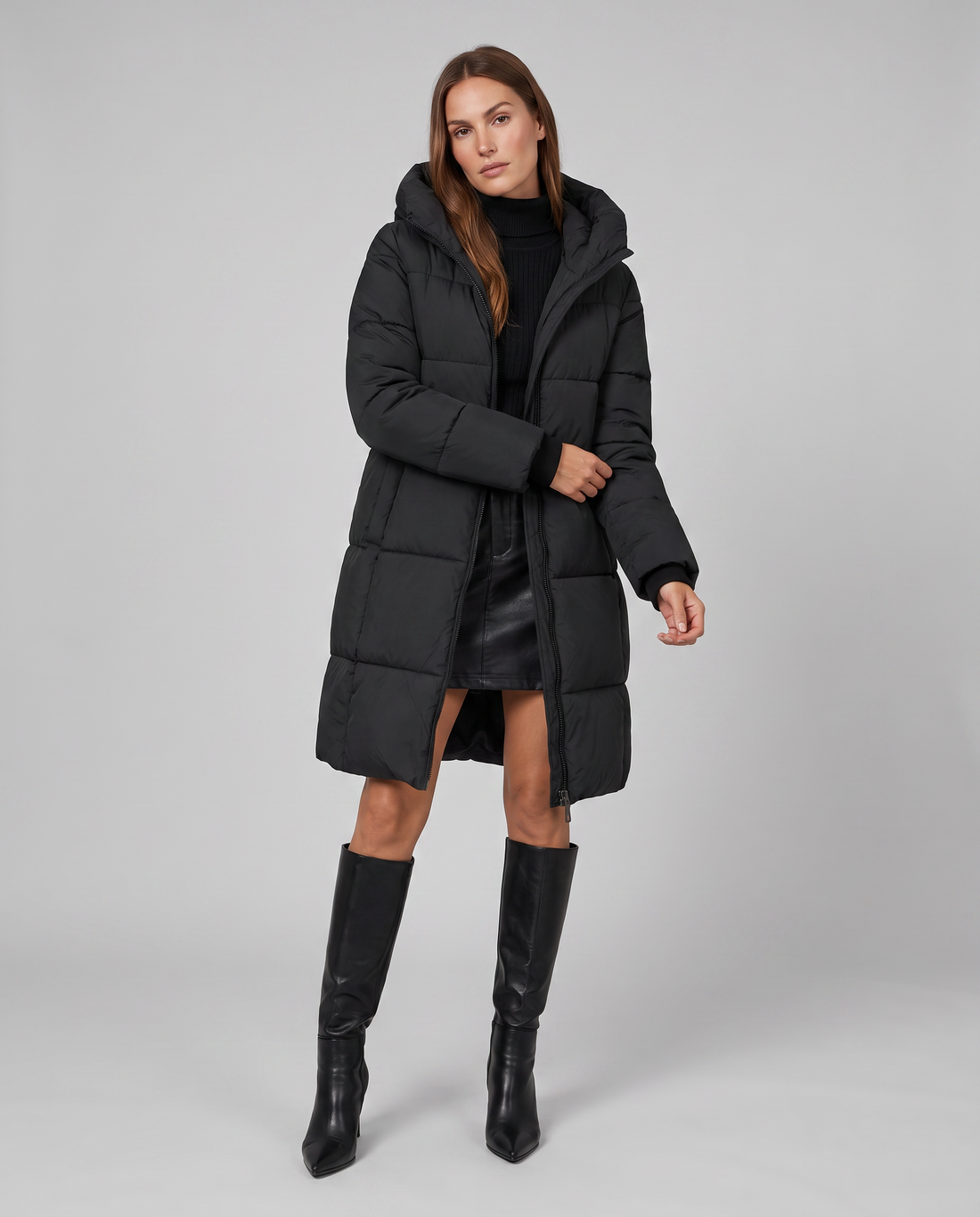 Woman wearing black hooded quilted puffer coat with long sleeves, paired with black leather skirt and knee-high black boots
