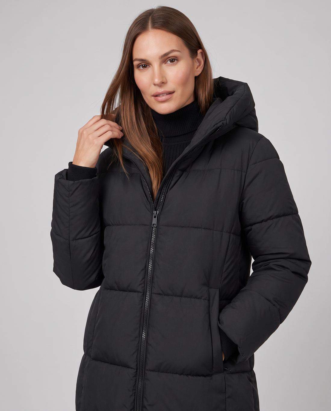 Woman wearing black quilted puffer coat with hood and black turtleneck, hand near shoulder against neutral background