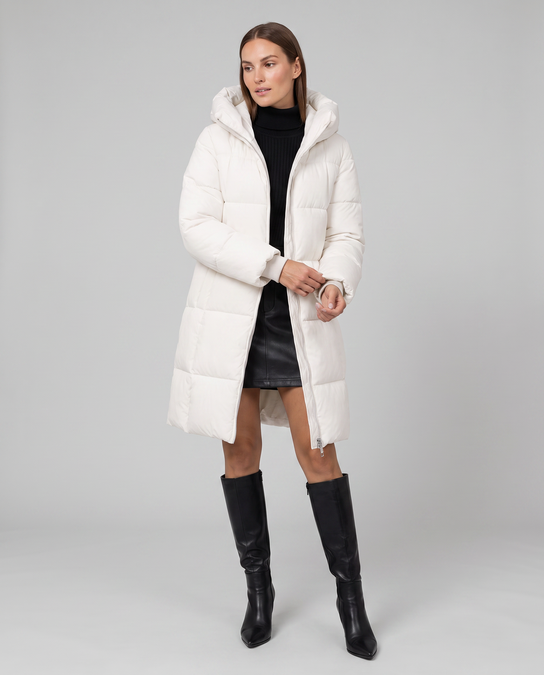 Woman wearing white quilted puffer coat with hood over black turtleneck and leather skirt, styled with black knee-high boots