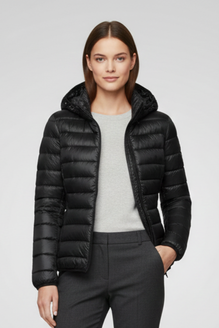 Woman wearing black quilted hooded zip jacket over gray shirt with black trousers, standing against white background