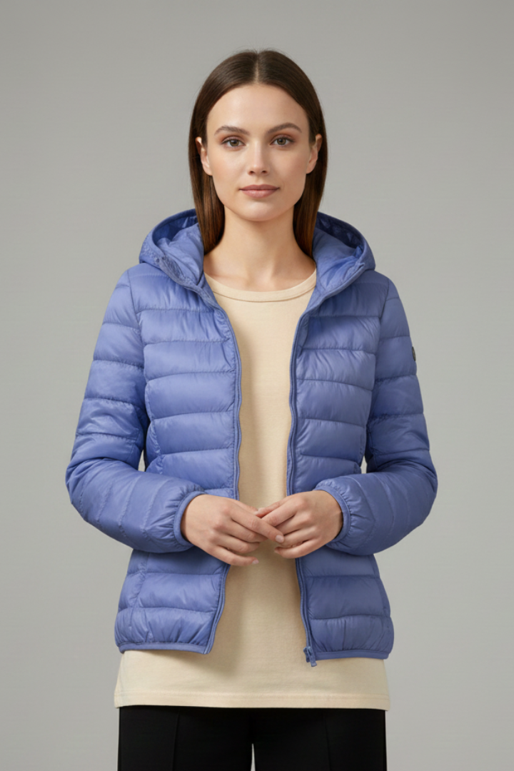 Woman wearing a blue quilted hooded zip-up jacket over a cream top and black pants against a gray background
