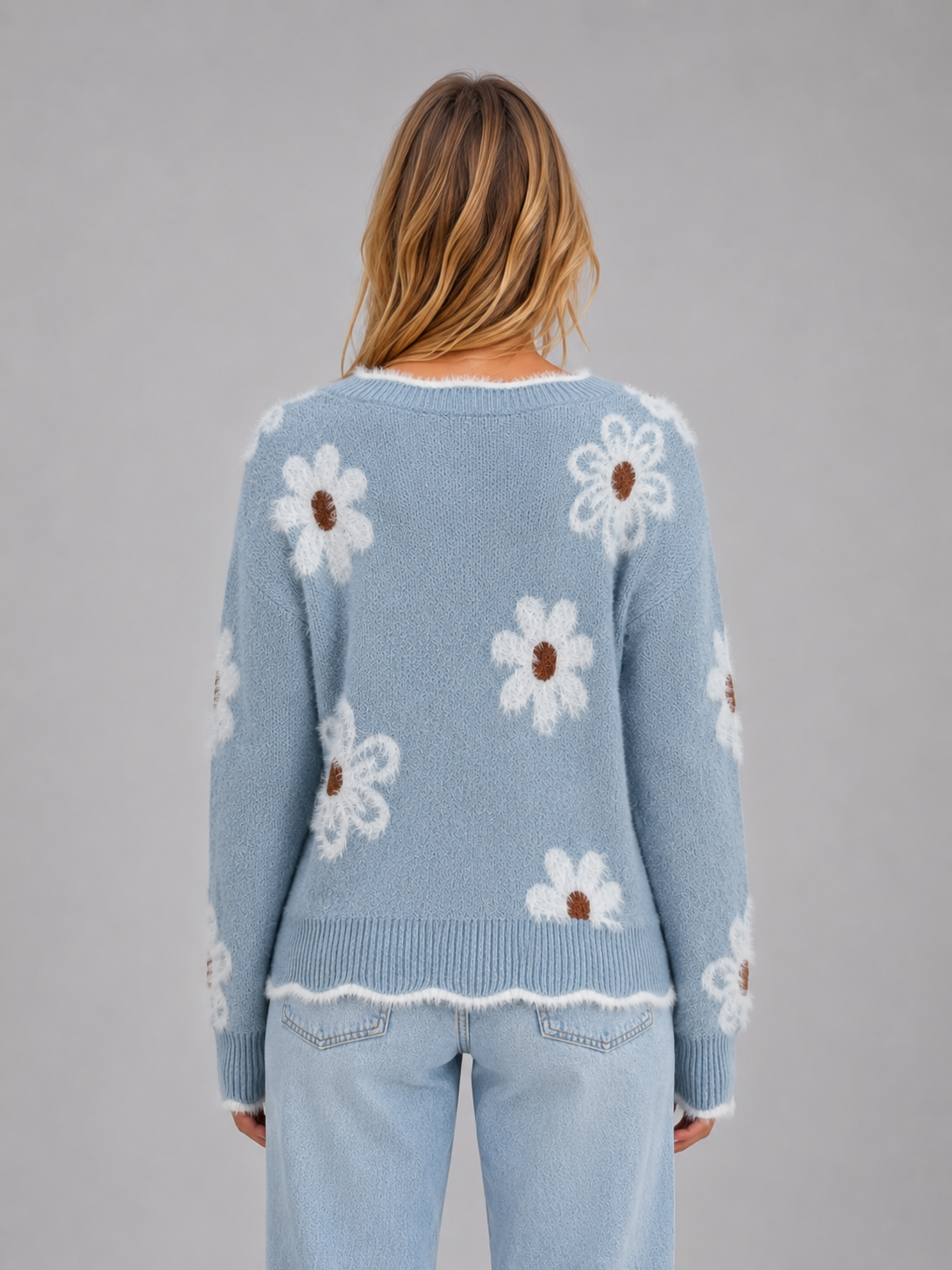Woman wearing light blue knit sweater with white daisy floral pattern and brown centers, paired with light wash jeans