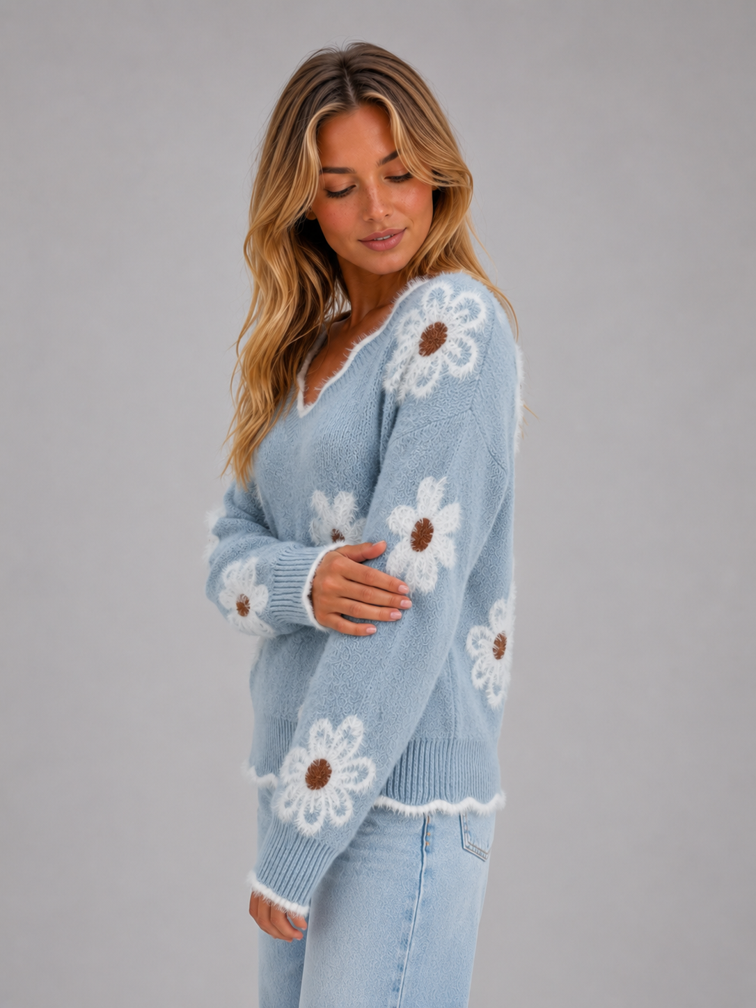 Woman wearing light blue soft knit v-neck pullover jumper with white daisy floral pattern and brown centers, paired with light blue jeans