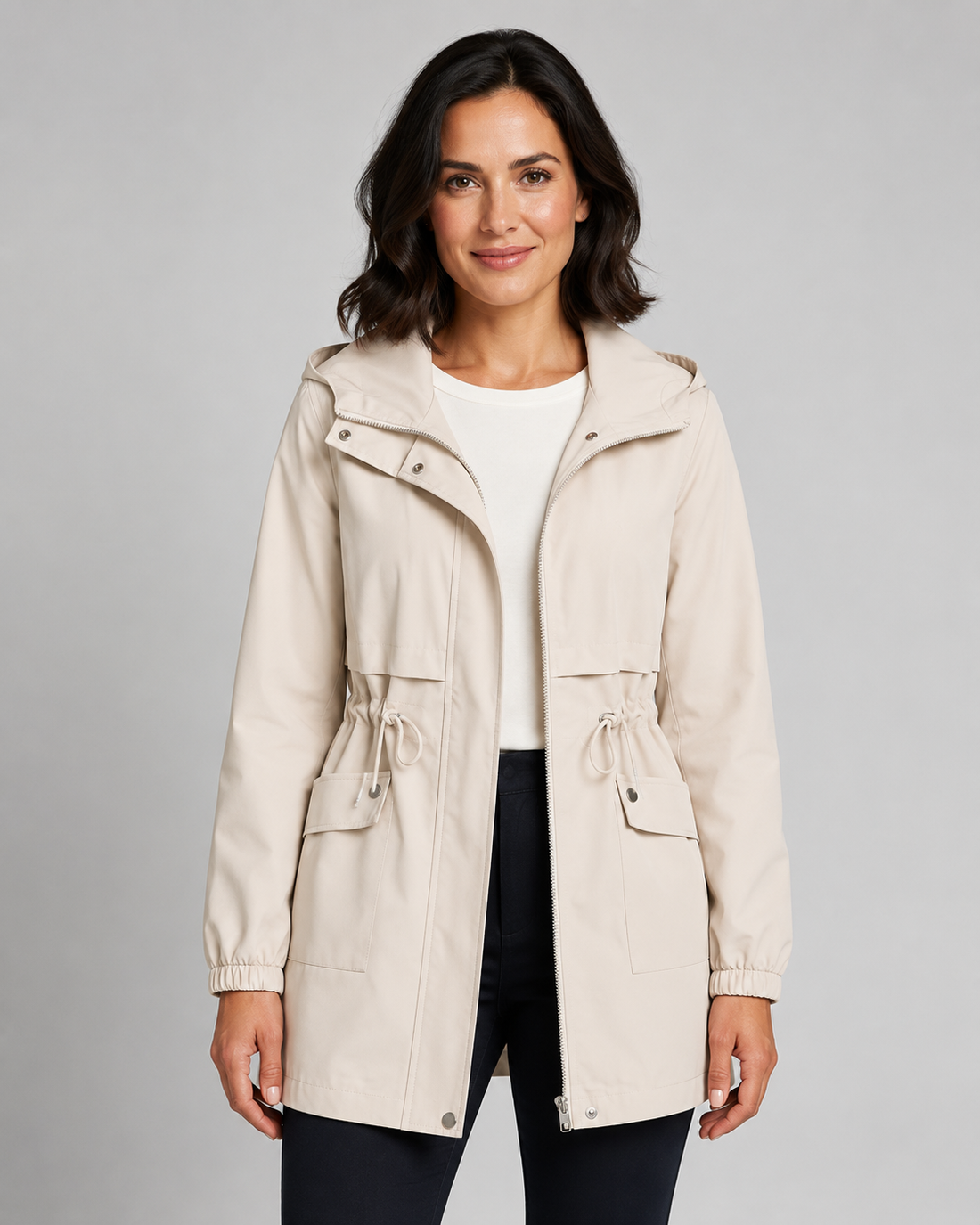 Woman wearing beige hooded trench coat with drawstring waist over white shirt and black pants, front view on gray background