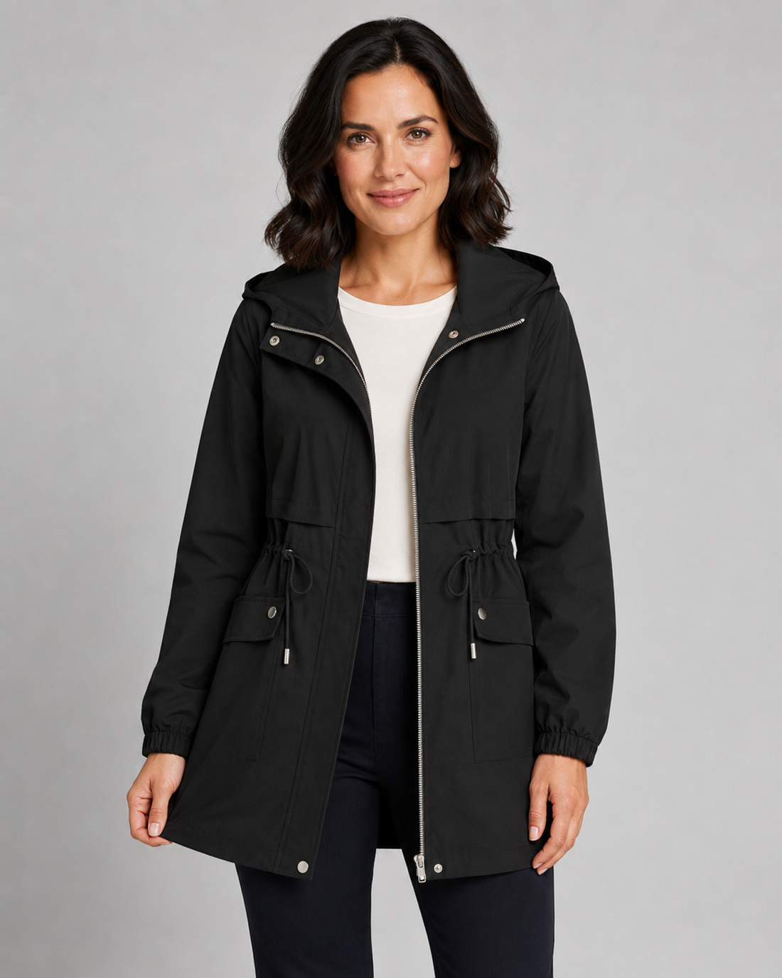 Woman wearing black hooded trench coat with drawstring waist, white top, and dark pants, standing against gray background