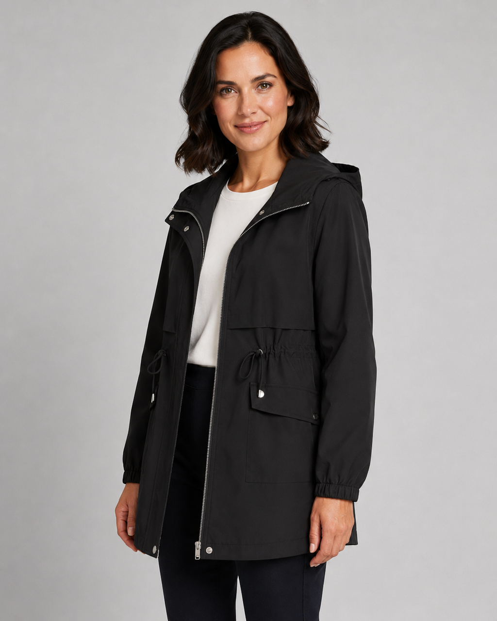 Woman wearing black hooded trench coat with drawstring waist over white shirt and black pants against gray background