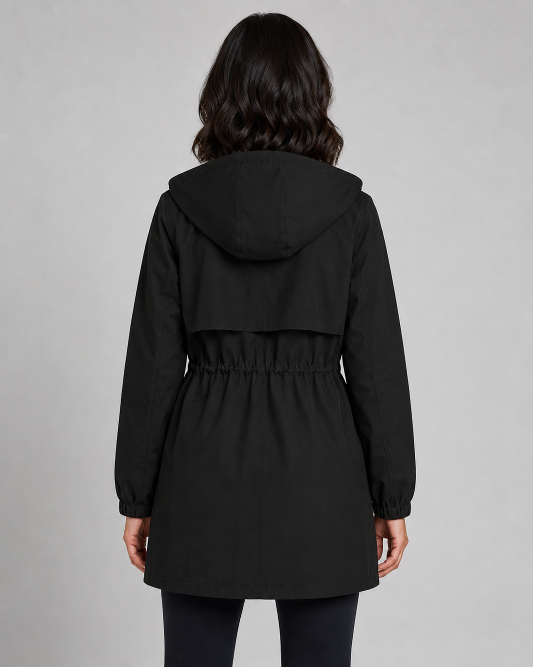 Woman wearing black trench coat with hood and drawstring waist, shown from behind against gray background
