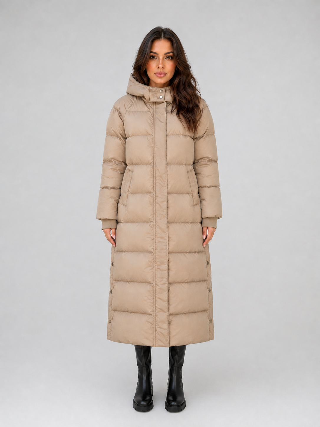 Woman wearing beige hooded padded puffer coat with quilted sections and black boots on white background