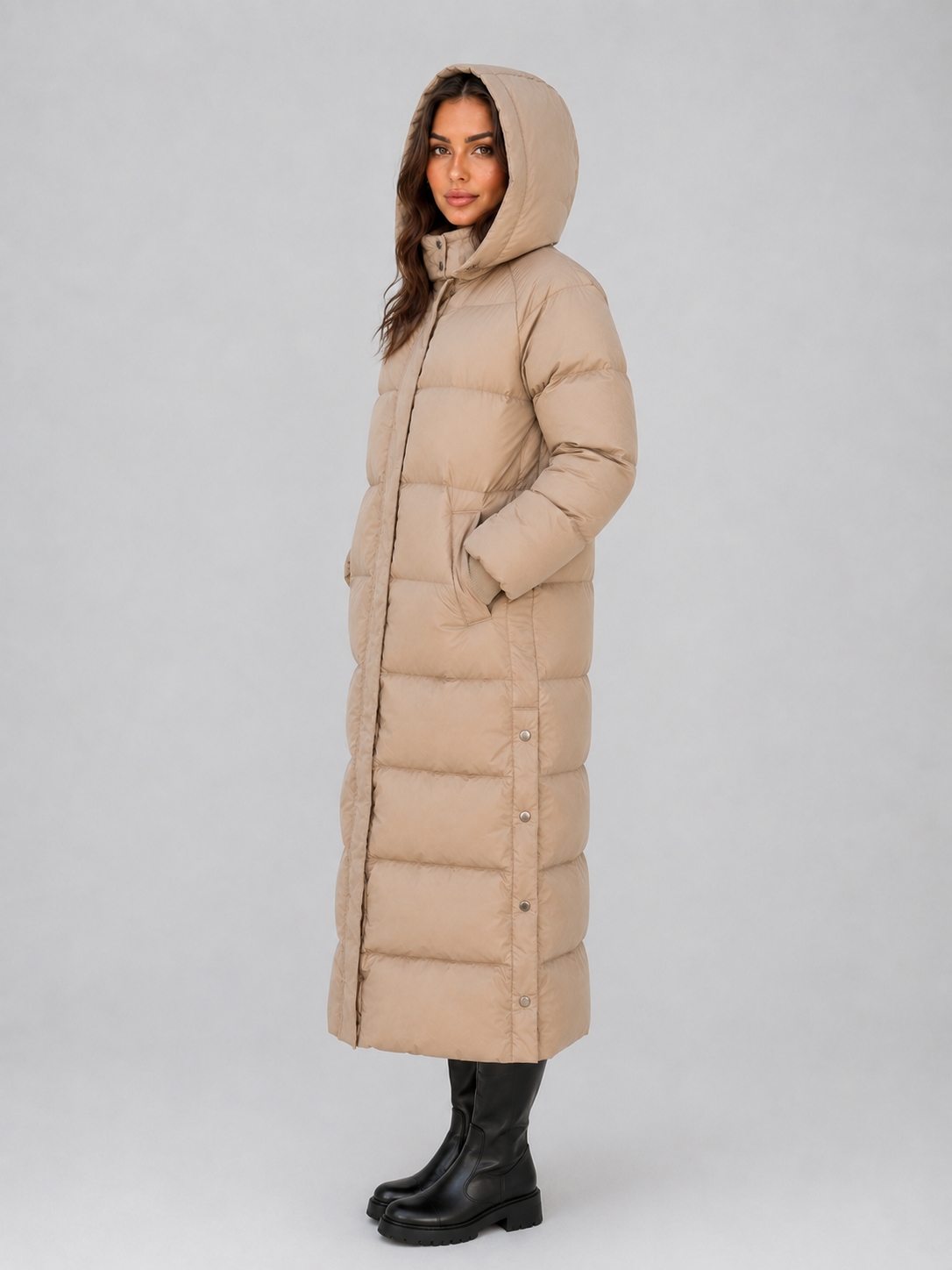 Woman wearing beige hooded padded long puffer winter coat with black boots, shown from side angle against white background