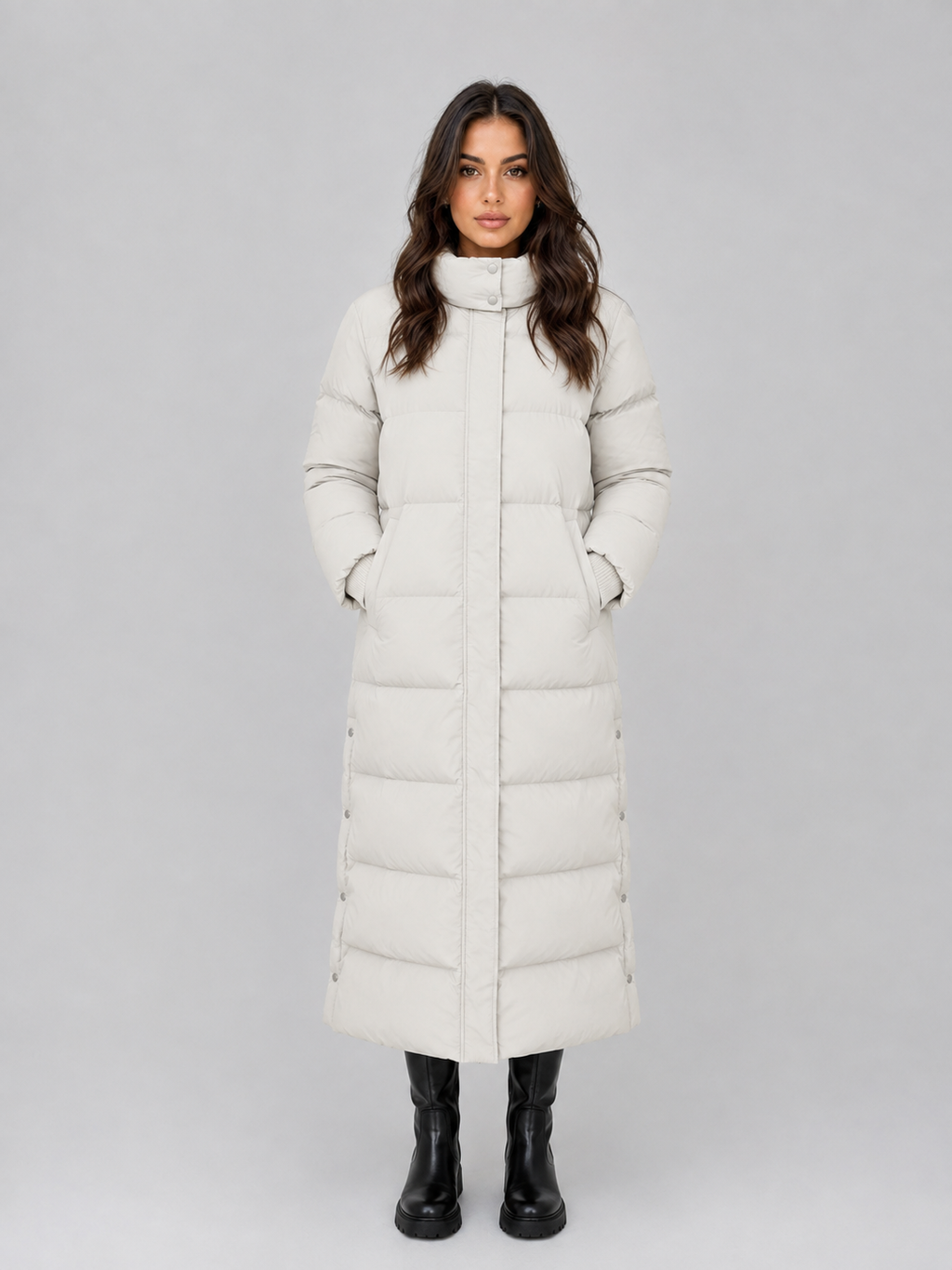 Woman wearing cream hooded padded long puffer winter coat with black boots, standing against gray background