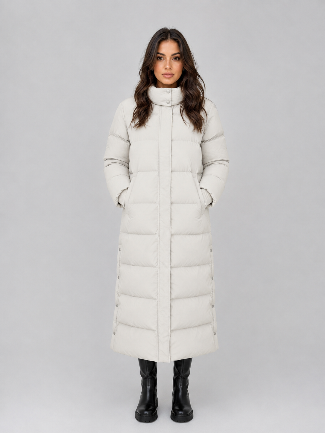 Woman wearing cream hooded padded long puffer winter coat with black boots, standing against gray background