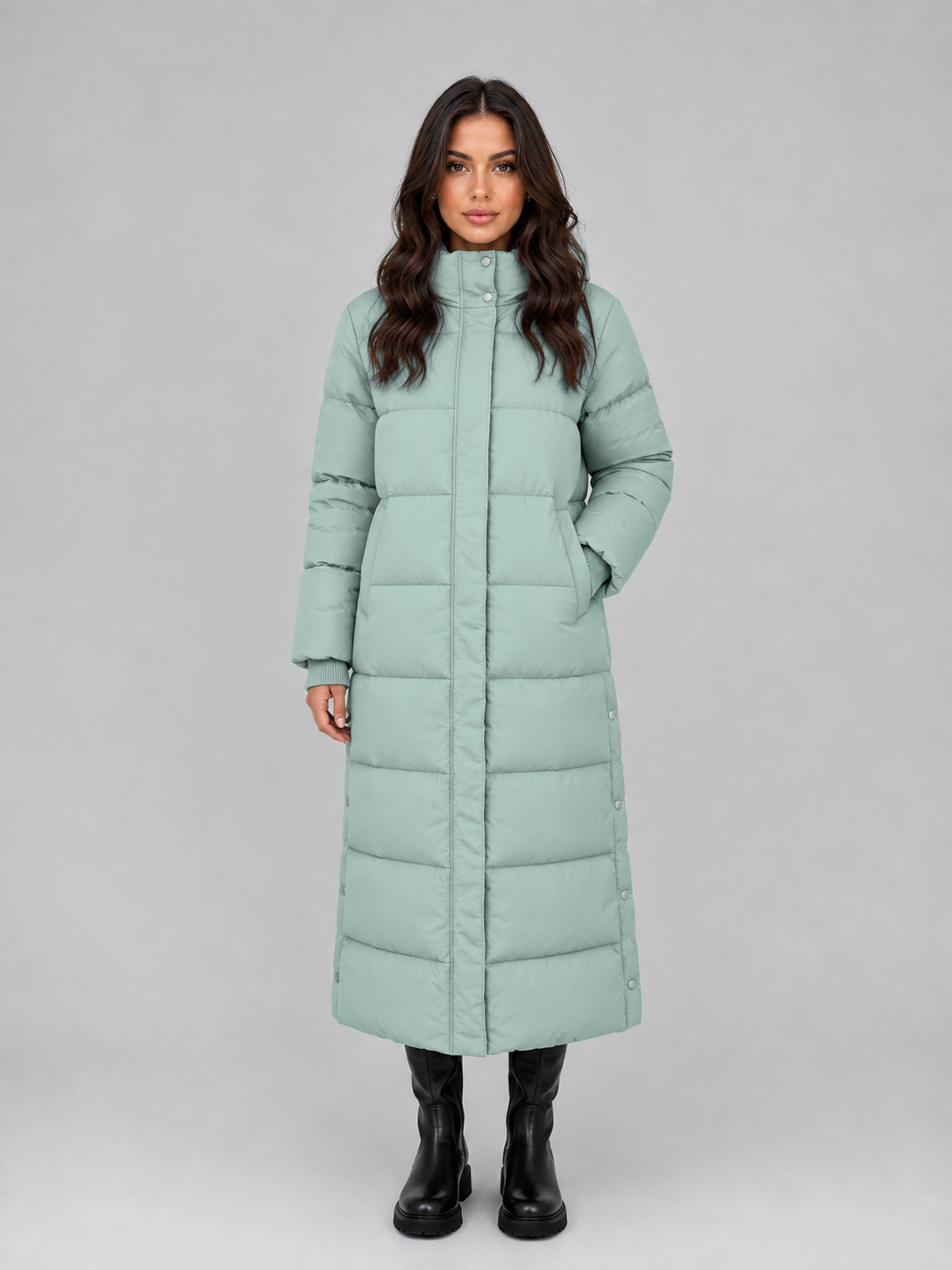 Woman wearing a light green quilted puffer coat with hood and button front, paired with black boots against a gray background