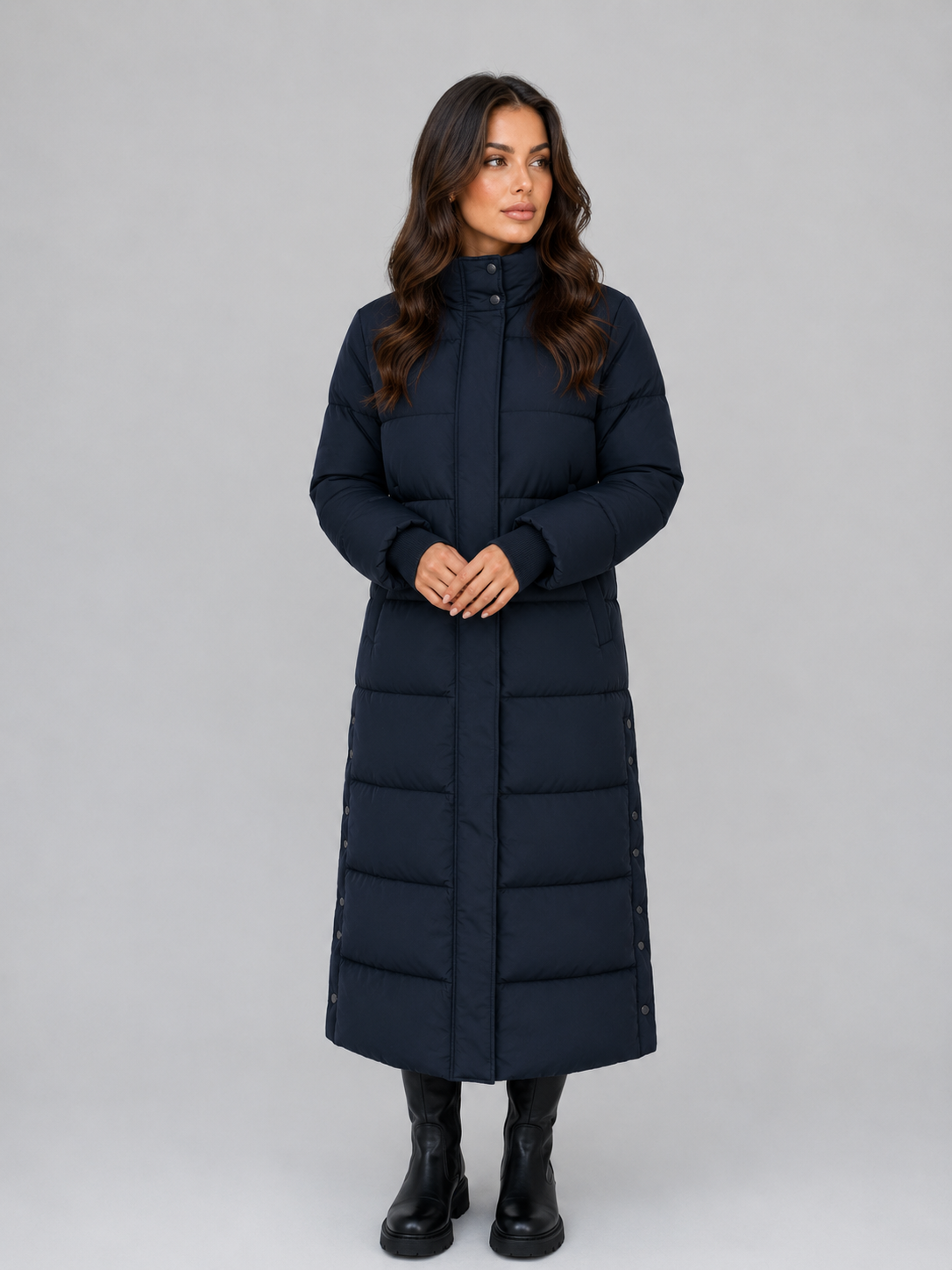 Woman wearing black hooded padded long puffer winter coat with hands clasped, paired with black boots against gray background