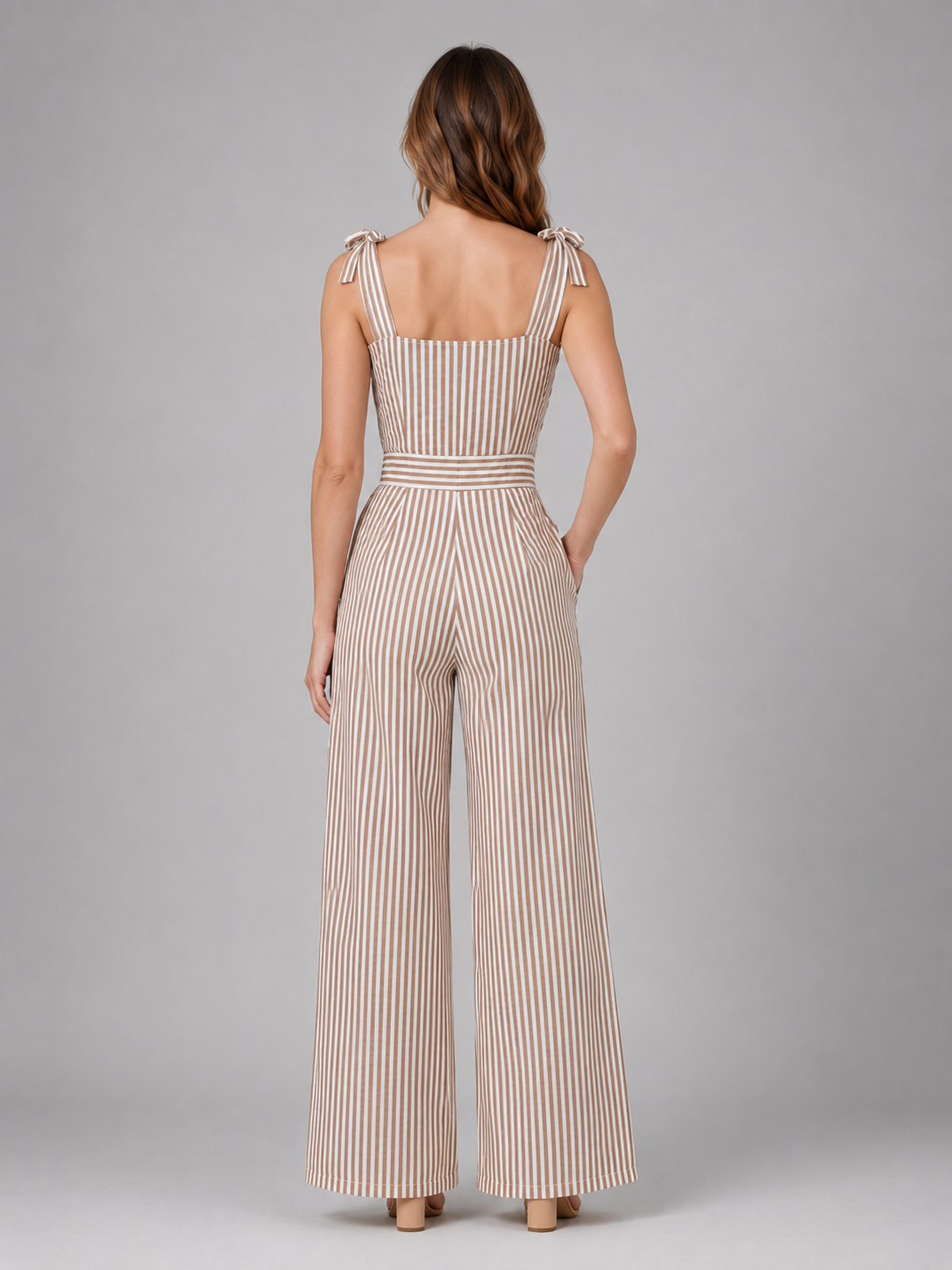 Woman wearing beige and white striped wide leg jumpsuit with tie shoulder straps, shown from back view on gray background