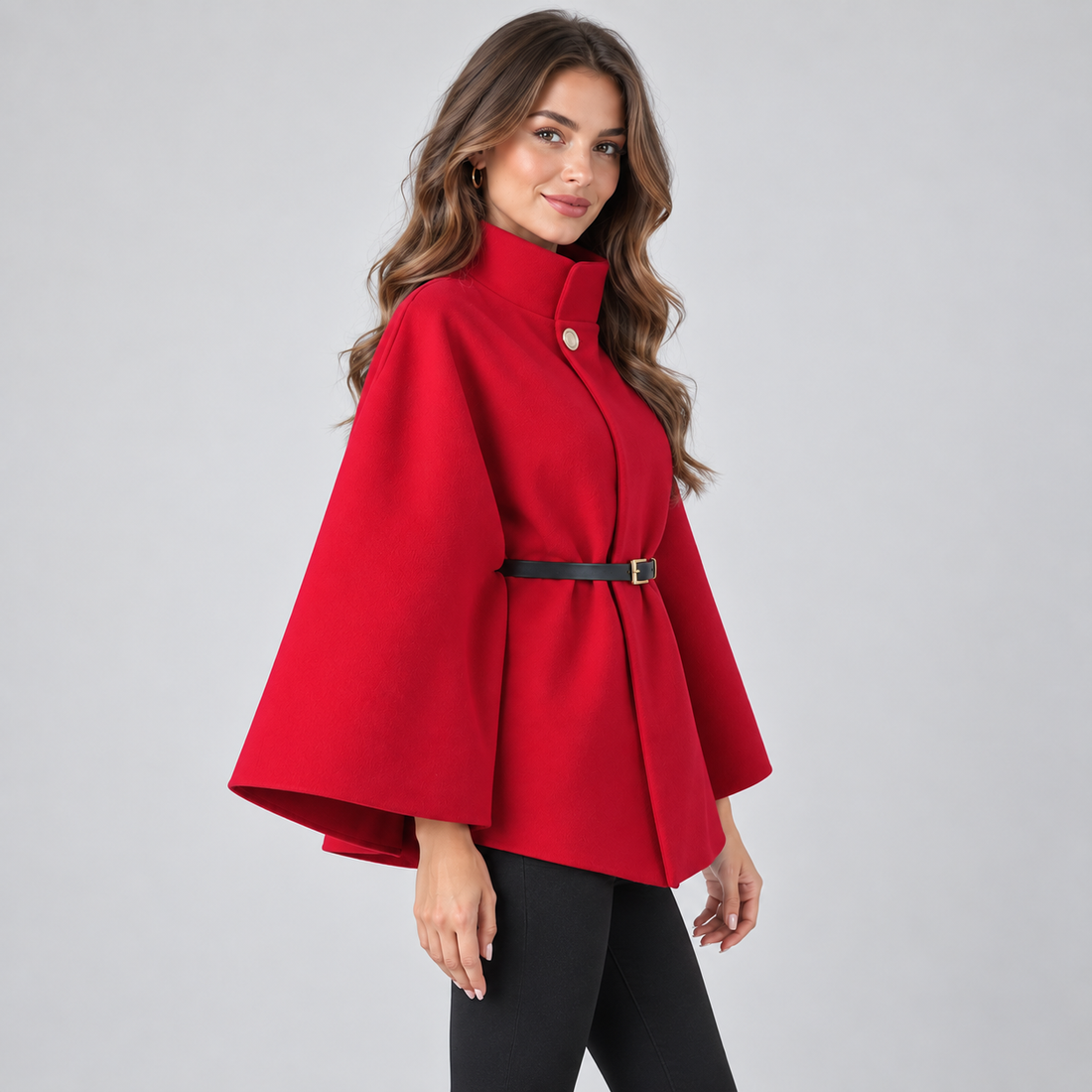 Woman wearing red high neck belted cape coat with black pants, standing against gray background