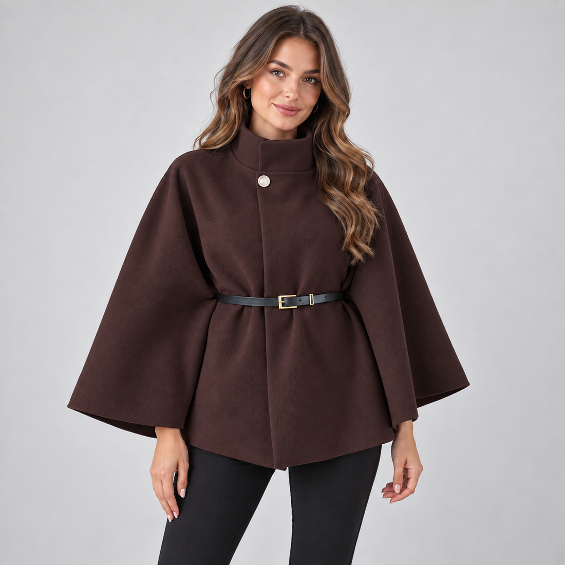 Woman wearing a dark brown high neck belted cape coat with black pants and gold hoop earrings, standing against a light gray background