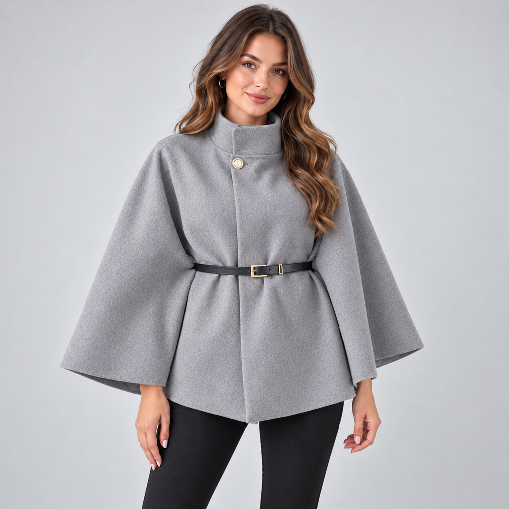 Woman wearing gray high neck belted cape coat with black pants and gold hoop earrings against light background