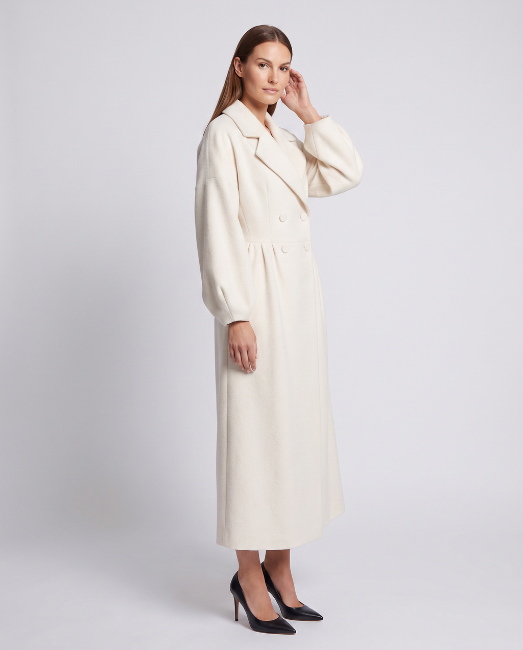 Woman wearing white double breasted maxi coat with long sleeves and black pointed toe heels, standing against light gray background